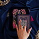 An image of the blue Serpents & Swords eReader decor bundle. The set features an eReader sleeve, eReader sticker insert and Clingie (think an enamel pop socket!) all in the same navy colour with bright ink, blue and teal detailing alongside gold foil and embroidery. The intricate design features motifs of crowns, swords, goblets and eyes. The eReader case features a wrist strap and bright pink tassel.