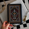 The Serpents & Swords eReader sticker insert is stuck to a kindle that is held over a classic book on a chessboard. The sticker features snake, crown and sword motifs and is black with gold, red and white detailing. The kindle also has the gold Gilded Heir dust plug, which features a gold crown motif.