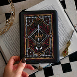 The Serpents & Swords eReader sticker insert is stuck to a kindle that is held over a classic book on a chessboard. The sticker features snake, crown and sword motifs and is black with gold, red and white detailing. The kindle also has the gold Gilded Heir dust plug, which features a gold crown motif.