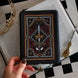 The Serpents & Swords eReader sticker insert is stuck to a kindle that is held over a classic book on a chessboard. The sticker features snake, crown and sword motifs and is black with gold, red and white detailing. The kindle also has the gold Gilded Heir dust plug, which features a gold crown motif.