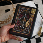 The Serpents & Swords eReader sticker insert is stuck to a kindle that is held over a classic book on a chessboard. The sticker features snake, crown and sword motifs and is black with gold, red and white detailing. The kindle also has the gold Gilded Heir dust plug, which features a gold crown motif. Attached to the kindle is also the Serpents & Sword charmlet in gold, attached via a charm anchor.