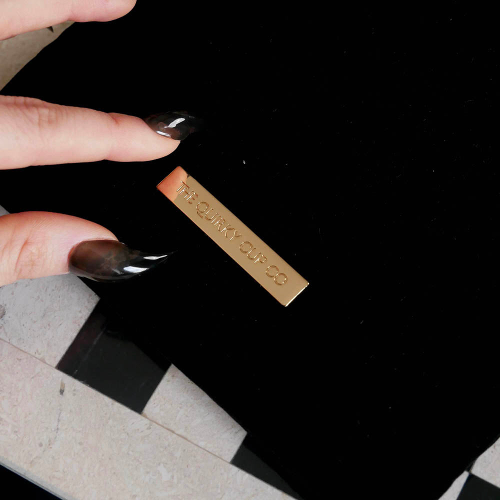 The Serpents & Swords fantasy inspired eReader sleeve sits on a chessboard. The romantasy inspired velvet kindle case features snake, crown and sword embroidered motifs and is black with gold, red and white detailing. The case also features a wrist strap, red tassel and gold logo plate on the back.