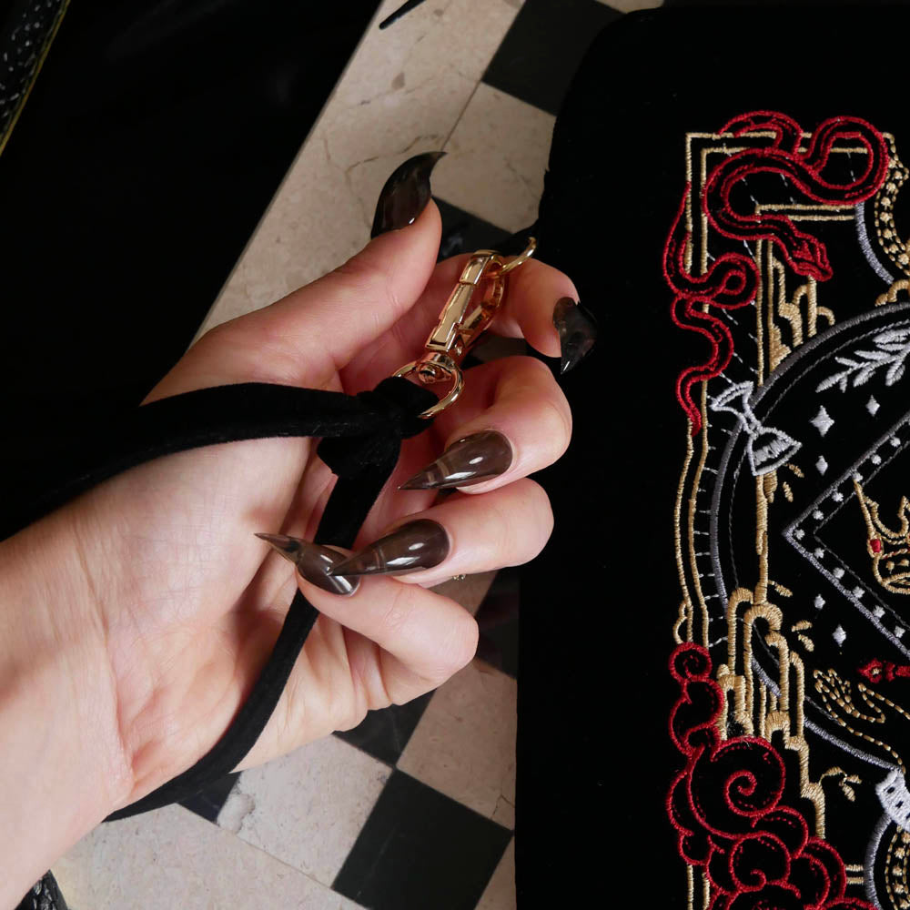 The Serpents & Swords fantasy inspired eReader sleeve sits on a chessboard on a black fabric background surrounded by black snakes. The romantasy inspired velvet kindle case features snake, crown and sword embroidered motifs and is black with gold, red and white detailing. The case also features a wrist strap and a red tassel.
