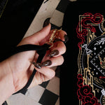 The Serpents & Swords fantasy inspired eReader sleeve sits on a chessboard on a black fabric background surrounded by black snakes. The romantasy inspired velvet kindle case features snake, crown and sword embroidered motifs and is black with gold, red and white detailing. The case also features a wrist strap and a red tassel.