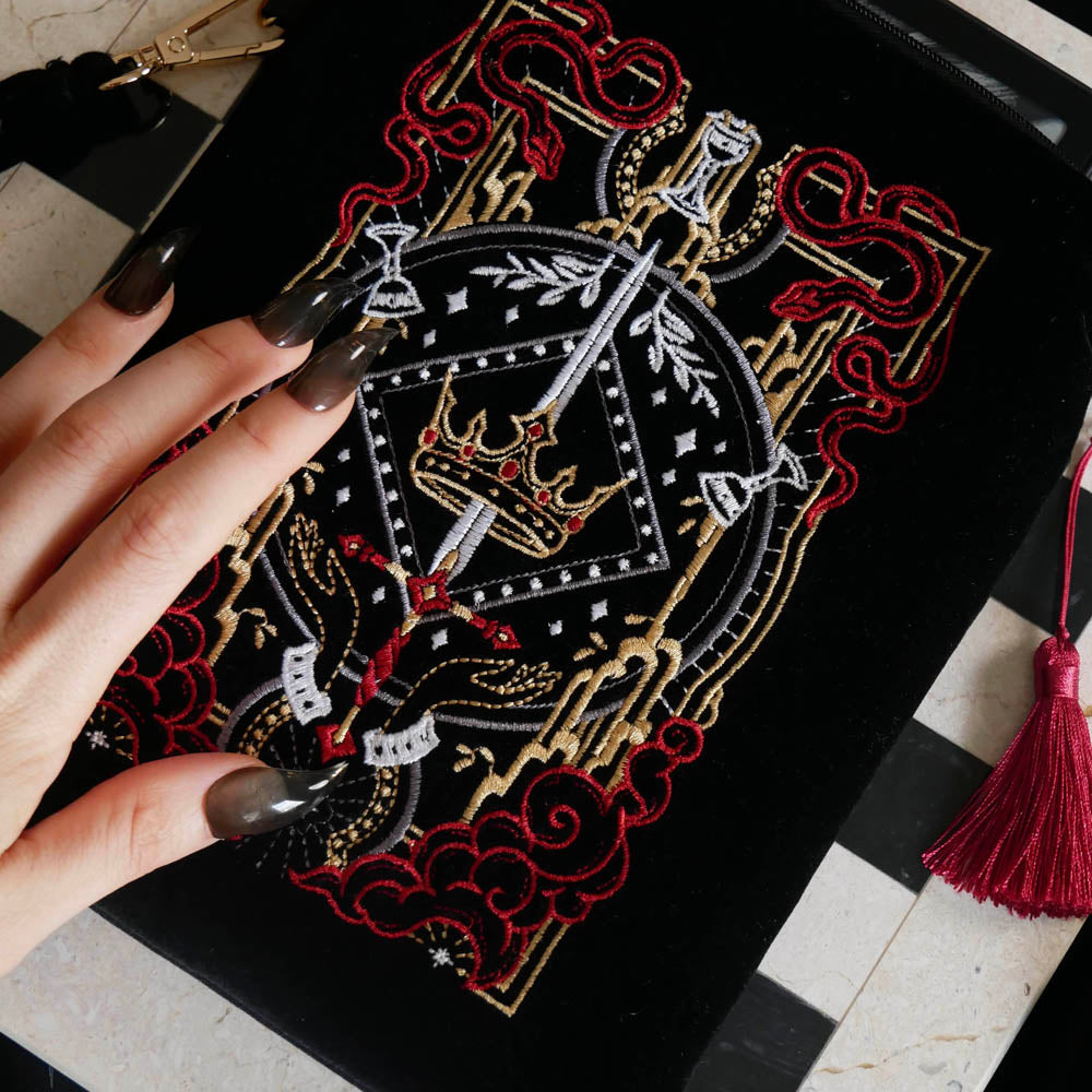 A detail of the Serpents & Swords eReader sleeve sits on a chessboard on a black fabric background surrounded by black snakes. The velvet kindle case features snake, crown and sword motifs and is black with gold, red and white detailing. he case also features a wrist strap and a red tassel.