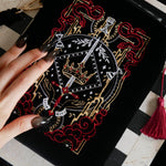 A detail of the Serpents & Swords eReader sleeve sits on a chessboard on a black fabric background surrounded by black snakes. The velvet kindle case features snake, crown and sword motifs and is black with gold, red and white detailing. he case also features a wrist strap and a red tassel.