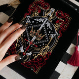 A detail of the Serpents & Swords eReader sleeve sits on a chessboard on a black fabric background surrounded by black snakes. The velvet kindle case features snake, crown and sword motifs and is black with gold, red and white detailing. he case also features a wrist strap and a red tassel.