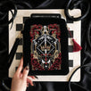 The Serpents & Swords eReader sleeve sits on a chessboard on a black fabric background surrounded by black snakes. The velvet kindle case features snake, crown and sword motifs and is black with gold, red and white detailing. he case also features a wrist strap and a red tassel.