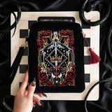 The Serpents & Swords eReader sleeve sits on a chessboard on a black fabric background surrounded by black snakes. The velvet kindle case features snake, crown and sword motifs and is black with gold, red and white detailing. he case also features a wrist strap and a red tassel.