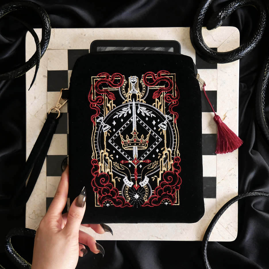 The Serpents & Swords eReader sleeve sits on a chessboard on a black fabric background surrounded by black snakes. The velvet kindle case features snake, crown and sword motifs and is black with gold, red and white detailing. he case also features a wrist strap and a red tassel.