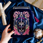 The Blue Serpents & Swords E-Reader Sleeve. On a blue fabric background, a hand holds the embroidered kindle case with an e-reader sticking out the top. The case features a pink tassel and pink, gold and blue toned embroidery showing a sword, crown and snake motif.