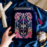 The Blue Serpents & Swords E-Reader Sleeve. On a blue fabric background, a hand holds the embroidered kindle case with an e-reader sticking out the top. The case features a pink tassel and pink, gold and blue toned embroidery showing a sword, crown and snake motif.