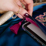A close up of the Blue Serpents & Swords E-Reader Sleeve. On a blue fabric background, a hand opens the kindle case to show the pink satin interior alongside the pink tassel and pink, gold and blue toned embroidery.