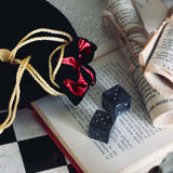 A black Quirky Cup Collective loot pouch with red interior and gold chord detailing sits on an open book and as chessboard, two black dice in the foreground.