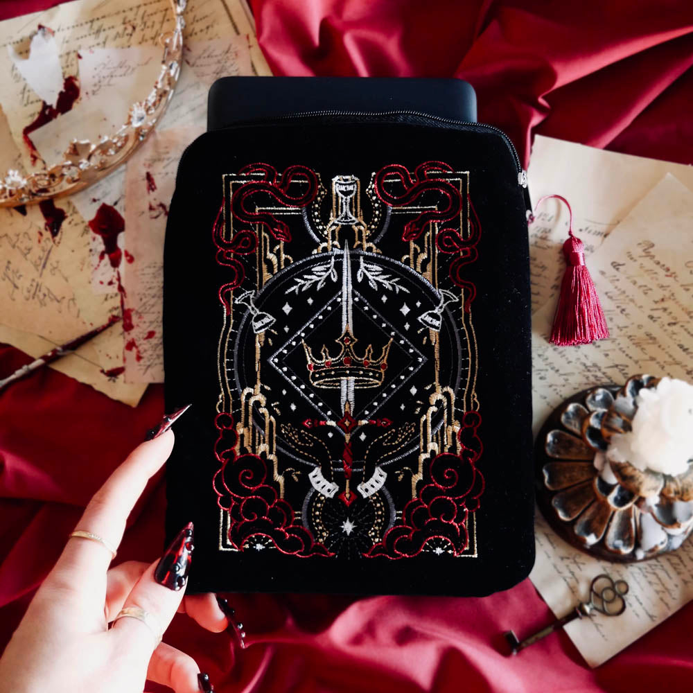 The black Serpents & Swords e-reader sleeve sits on a red fabric background surrounded by a crown, handwritten notes and a candle for a  romantasy aesthetic. The e-reader sleeve features intricate  embroidery of a snake, crown, sword and goblet motif in shades of red, white and gold. The book sleeve also has a red tassle.