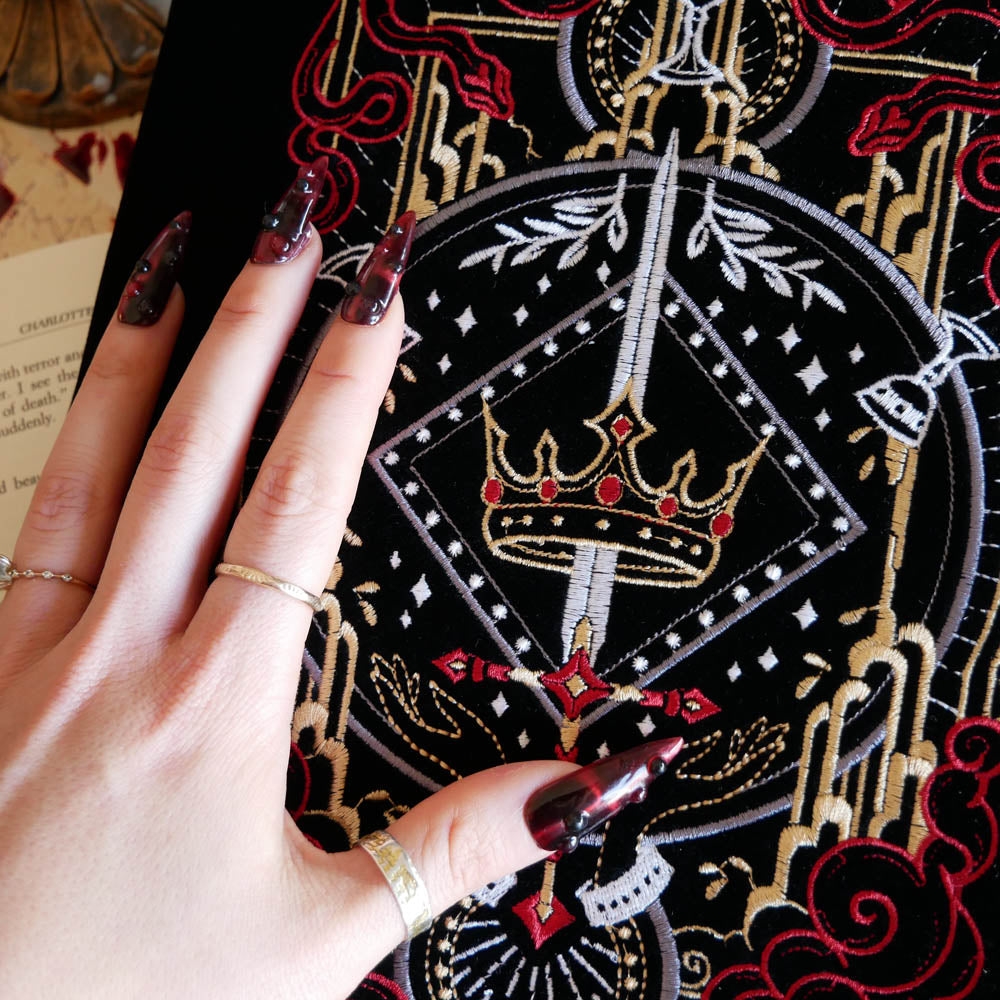 A detail of the  black Serpents & Swords book sleeve sees a hand with intricate nails show off the embroidery. The e-reader sleeve features intricate  embroidery of a snake, crown, sword and goblet motif in shades of red, white and gold. The book sleeve also has a red tassle.