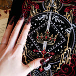 A detail of the  black Serpents & Swords book sleeve sees a hand with intricate nails show off the embroidery. The e-reader sleeve features intricate  embroidery of a snake, crown, sword and goblet motif in shades of red, white and gold. The book sleeve also has a red tassle.
