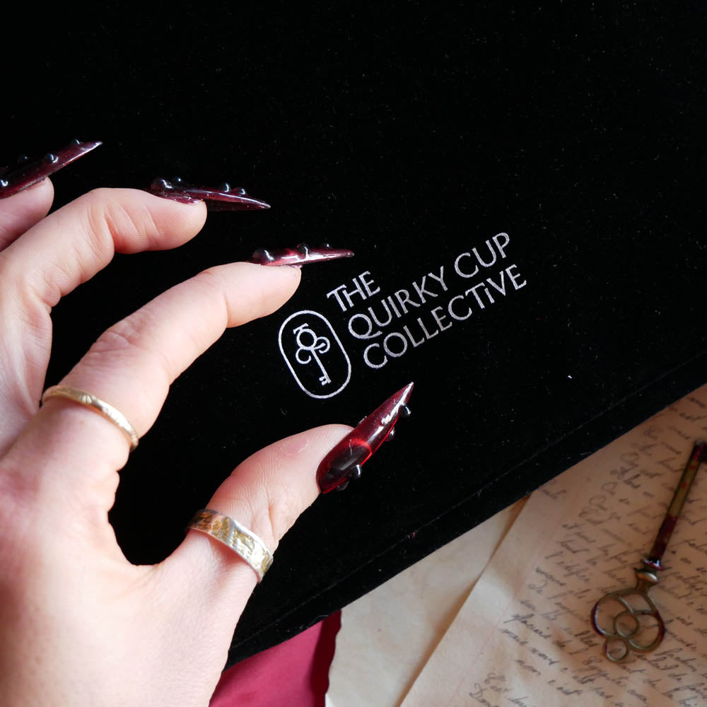 A close up of The Quirky Cup Collective logo on the black Serpents & Swords e-reader sleeve. The white logo features the key monogram