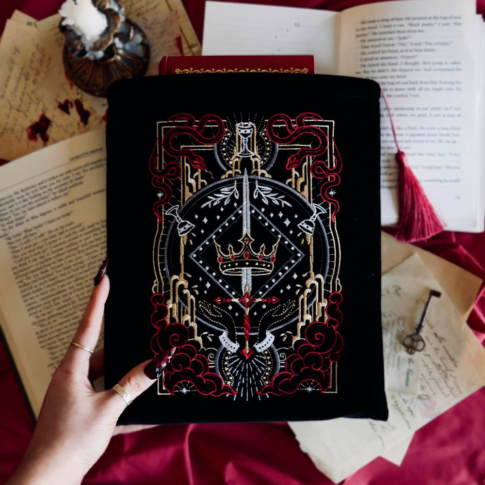 The black Serpents & Swords book sleeve sits on a red fabric background with surrounded by candles, books and handwritten letters for a romantasy aesthetic. The book sleeve features intricate embroidery of a snake, crown, sword and goblet motif in shades of red, white and gold. The book sleeve also has a red tassle.