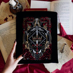 The black Serpents & Swords book sleeve sits on a red fabric background with surrounded by candles, books and handwritten letters for a romantasy aesthetic. The book sleeve features intricate embroidery of a snake, crown, sword and goblet motif in shades of red, white and gold. The book sleeve also has a red tassle.