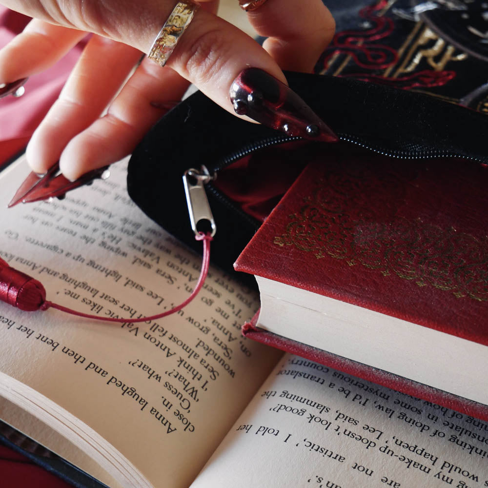 A detail of the black Serpents & Swords book sleeve sits on an open book background. The book sleeve features intricate embroidery of a snake, crown, sword and goblet motif in shades of red, white and gold. The book sleeve also has a red tassle and a red satin interior.