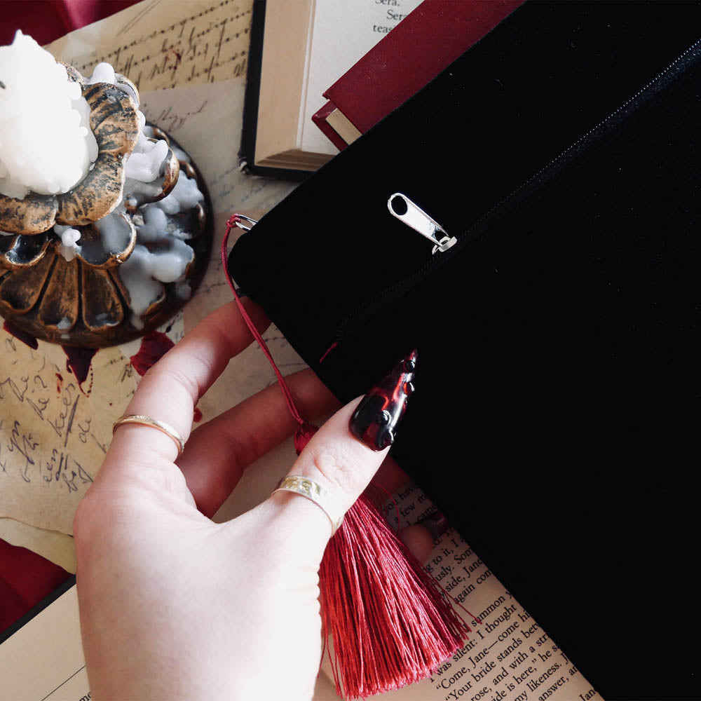A detail of the back of the black Serpents & Swords book sleeve sits on an open book background. The book sleeve features intricate embroidery of a snake, crown, sword and goblet motif in shades of red, white and gold. The book sleeve also has a red tassle and a red satin interior.