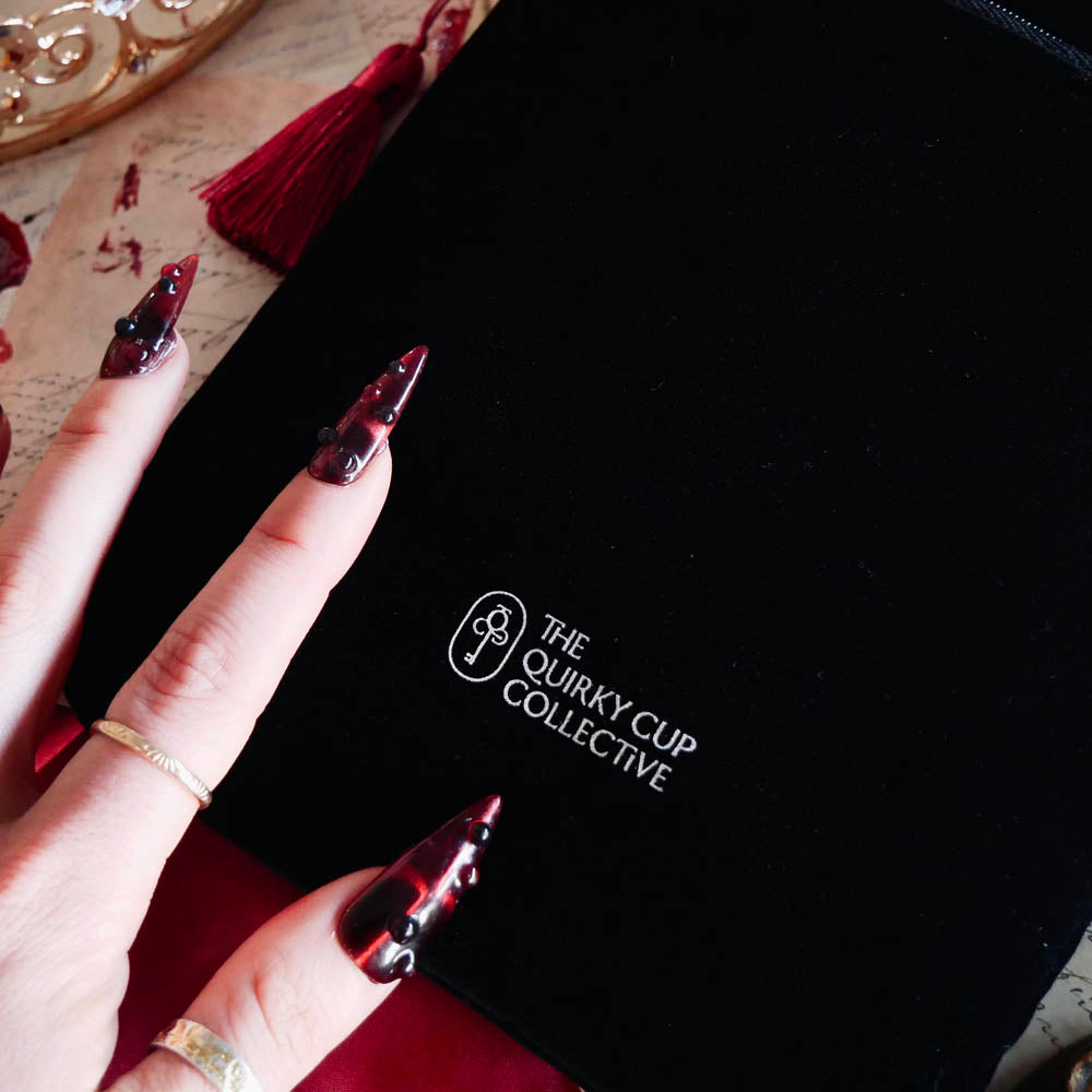 A close up of The Quirky Cup Collective logo on the black Serpents & Swords book sleeve. The white logo features the key monogram