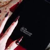 A close up of The Quirky Cup Collective logo on the black Serpents & Swords book sleeve. The white logo features the key monogram