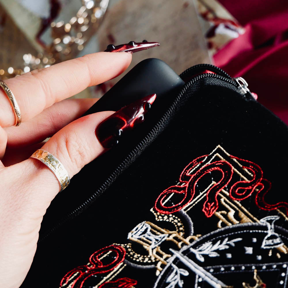 A detail of the black Serpents & Swords e-reader sleeve. A hand with intricate nails slides a kindle into the case. The e-reader sleeve features intricate  embroidery of a snake, crown, sword and goblet motif in shades of red, white and gold. The book sleeve also has a red tassle.