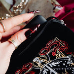 A detail of the black Serpents & Swords e-reader sleeve. A hand with intricate nails slides a kindle into the case. The e-reader sleeve features intricate  embroidery of a snake, crown, sword and goblet motif in shades of red, white and gold. The book sleeve also has a red tassle.