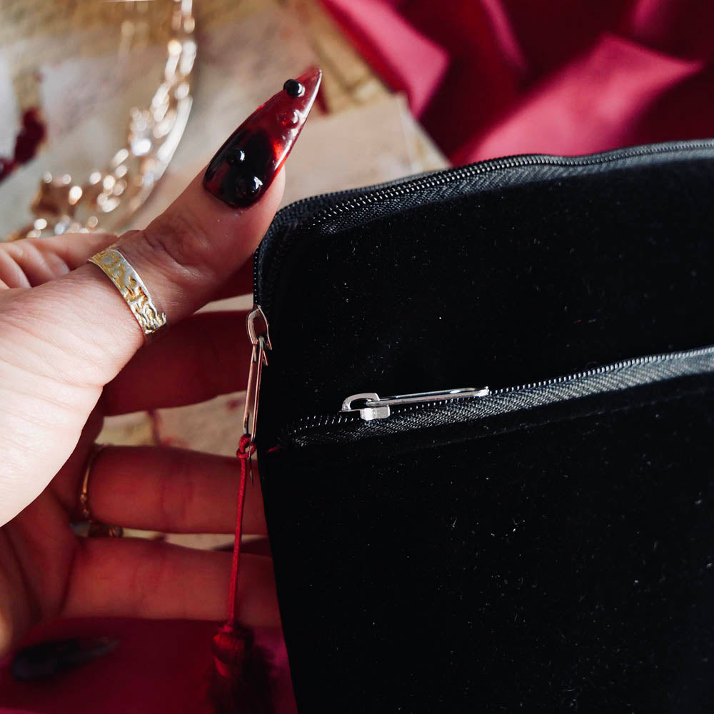 A close up of The Quirky Cup Collective black Serpents & Swords e-reader sleeve. A hand with intricate red nails shows off the velvet fabric and the red tassle on the zip
