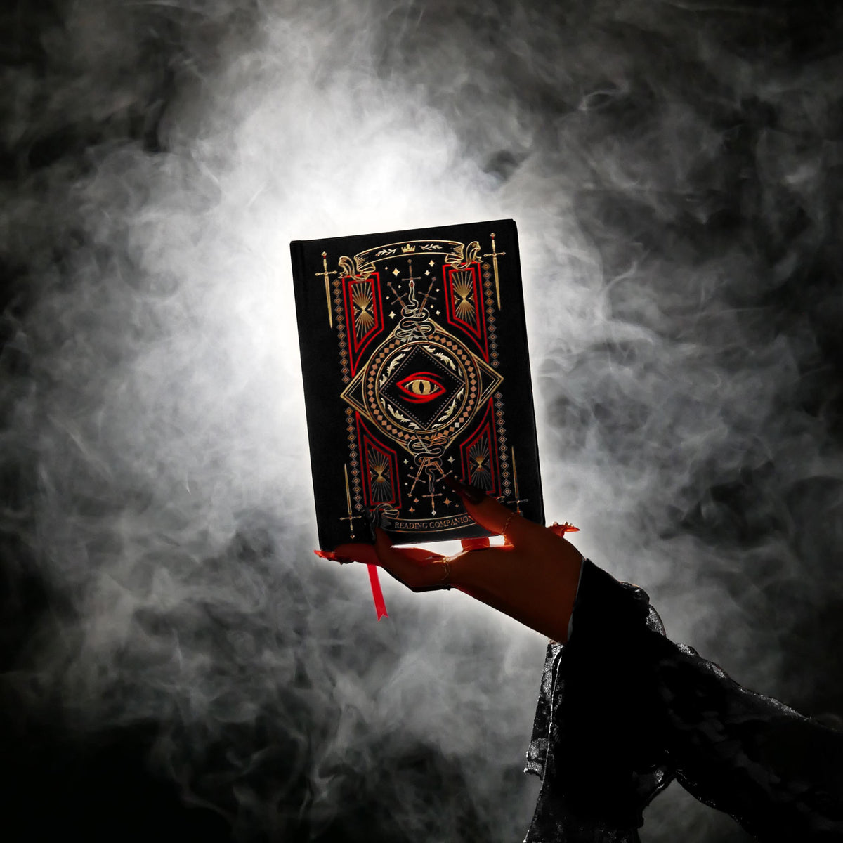 A moody fantasy inspired image from The Quirky Cup Collective showcasing the Serpents and Swords Collection. And intricately foiled journal (the Reading Companion) is held in a hand with long red nails in front of a smoky dark background. 