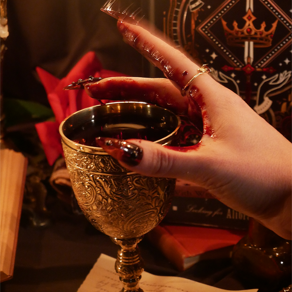 A moody image for The Quirky Cup Collective's Swords and Serpents collection. A hand with intricate red nails holds a goblet filled with blood in front of the fantasy book inspired stationery collection.