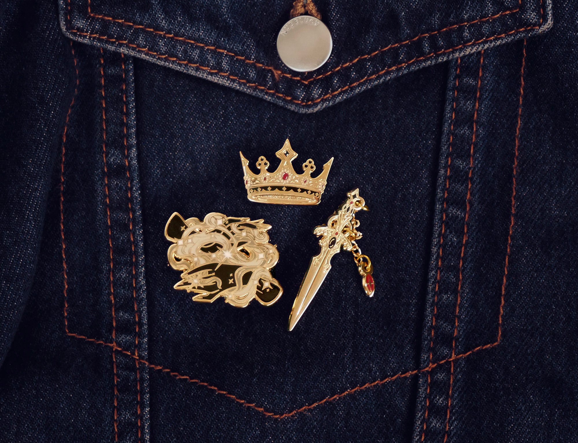 On a dark denim pocket, three of the Serpents & Swords enamel pins are shown. All gold, each design features black and red detailing. The Magic Weaver design shows two hands amidst swirling magic, The Gilder Heir design shows a small crown while the Dagger pin shows a small dagger with charm.
