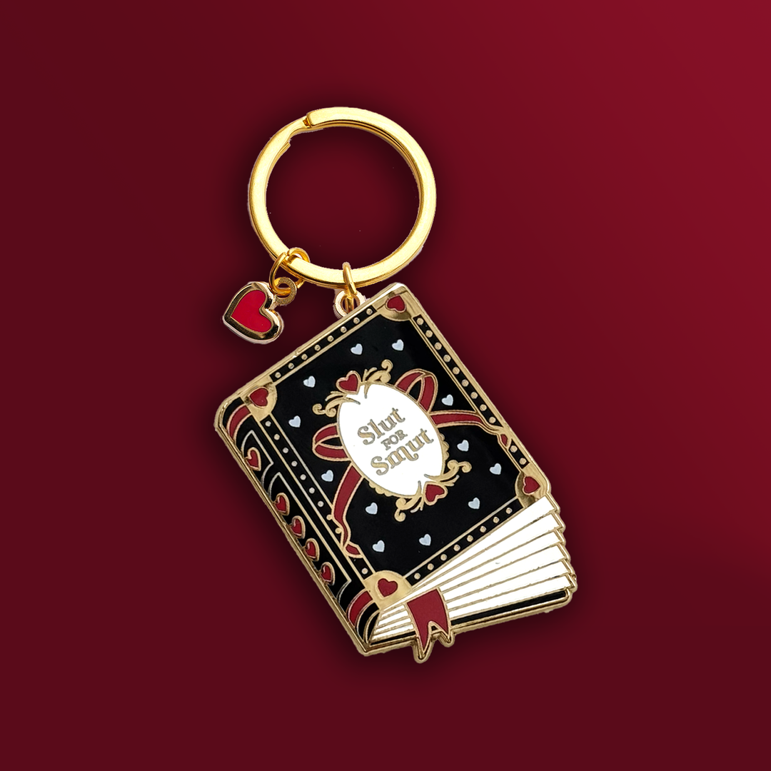 A keyring in the shape of book that has a black cover with red and metallic gold accents. The cover reads 'Slut for Smut' and features hearts and a bow. Keyring hardware is gold and has a heart charm with a red centre