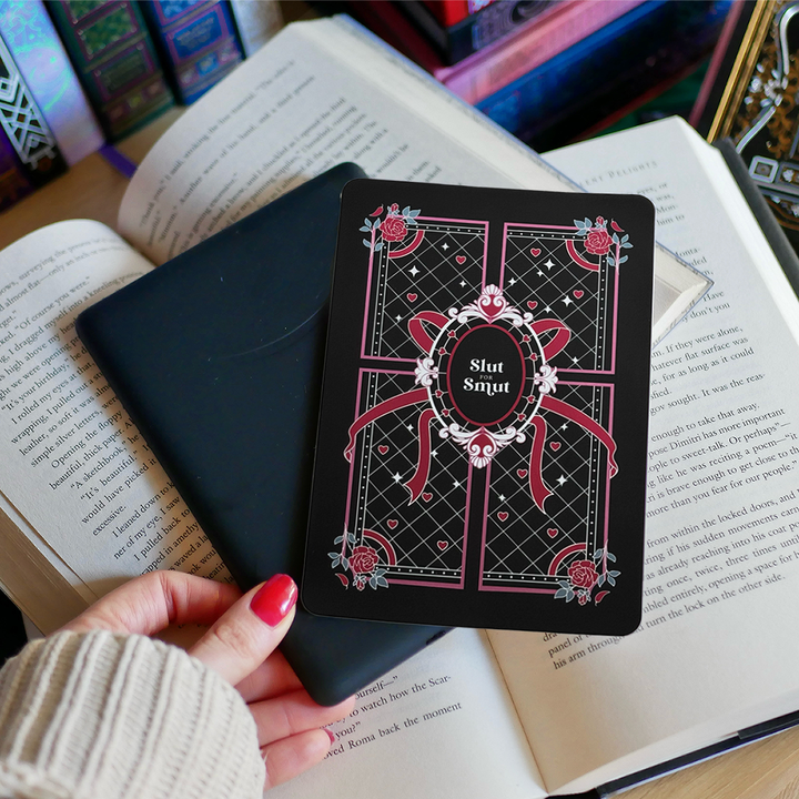 A hand holds the back of a kindle that sits on a bed of open books. On top of the kindle sits a sticker that reads 'slut for smut'. The sticker has black, red, pink and white accents that features hearts, roses and a ribbon on a quilted style pattern