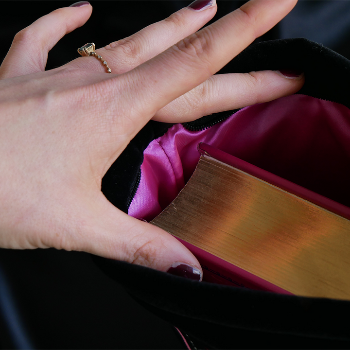 hand holding open a black velvet book sleeve with pink satin lining. Inside is a book with a pink cover with gold foil gilded edges
