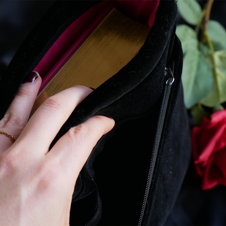 hand holding open a black velvet book sleeve with pink satin lining. Inside is a book with a pink cover with gold foil gilded edges

