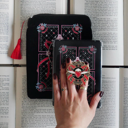 The Black Slut for Smut e-reader bundle set is shown on open book background and  includes a velvet e-reader case, enamel clingie and kindle sticker insert. The design is a black, pink and red feminine bow design, the clingie featuring a flying heart and bows in the same colour palette.