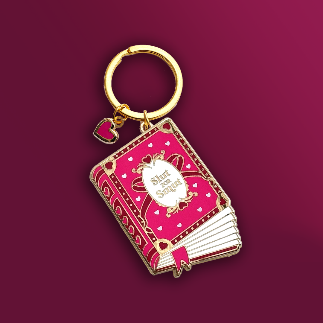 A keyring in the shape of book that has a pink cover with pink and metallic gold accents. The cover reads 'Slut for Smut' and features hearts and a bow. Keyring hardware is gold and has a heart charm with a pink centre