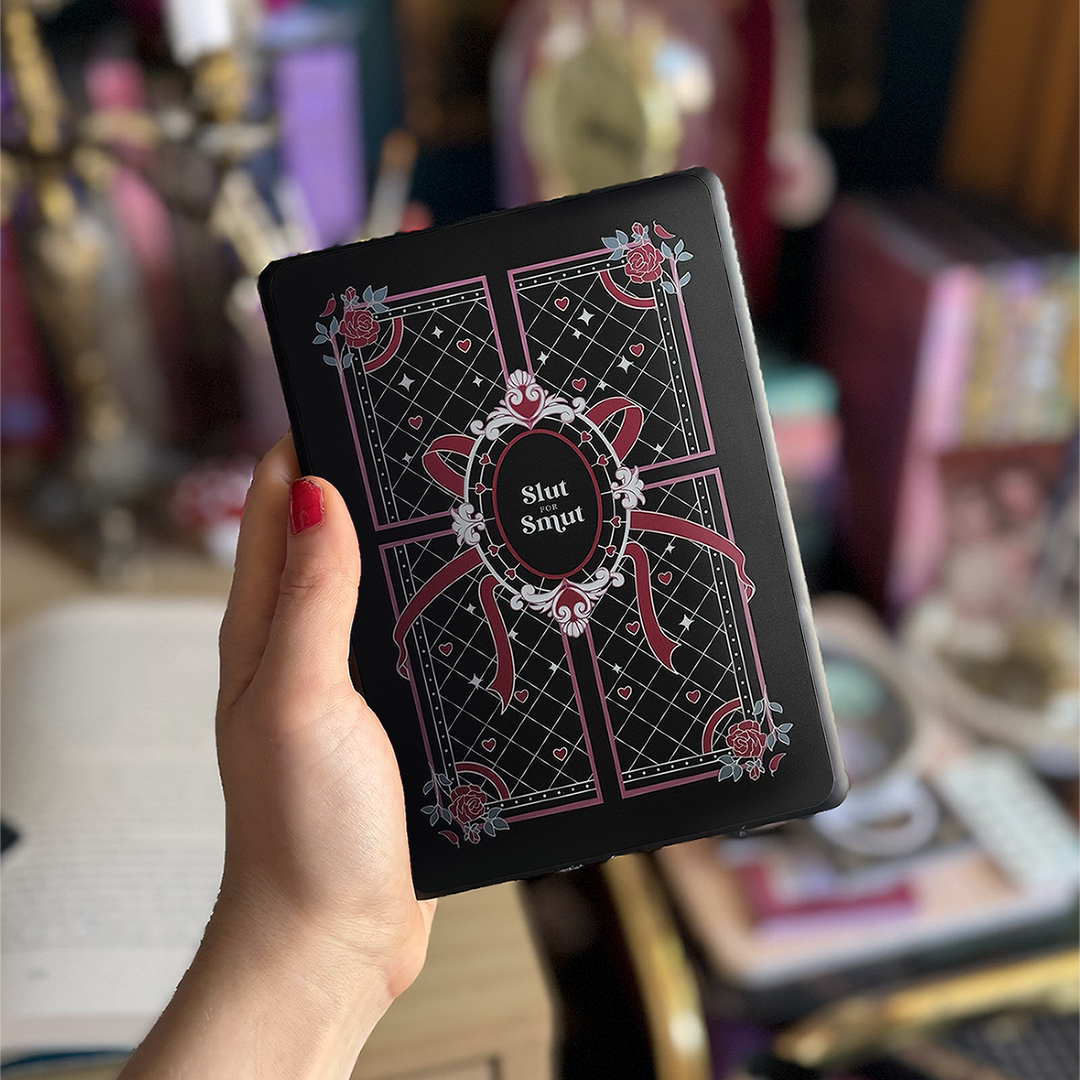 A hand holds up a kindle against a colourful office background. On the back of the kindle sits a sticker that reads 'slut for smut'. The sticker has black, pink white and teal accents that features hearts, roses and a ribbon on a quilted style pattern