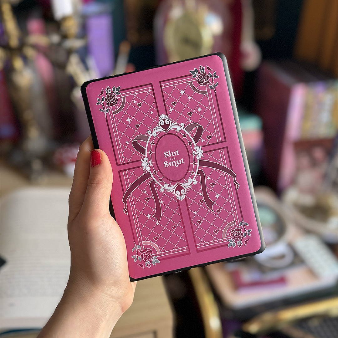 A hand holds up a kindle against a colourful office background. On the back of the kindle sits a sticker that reads 'slut for smut'. The sticker has pink, white and teal accents that features hearts, roses and a ribbon on a quilted style pattern