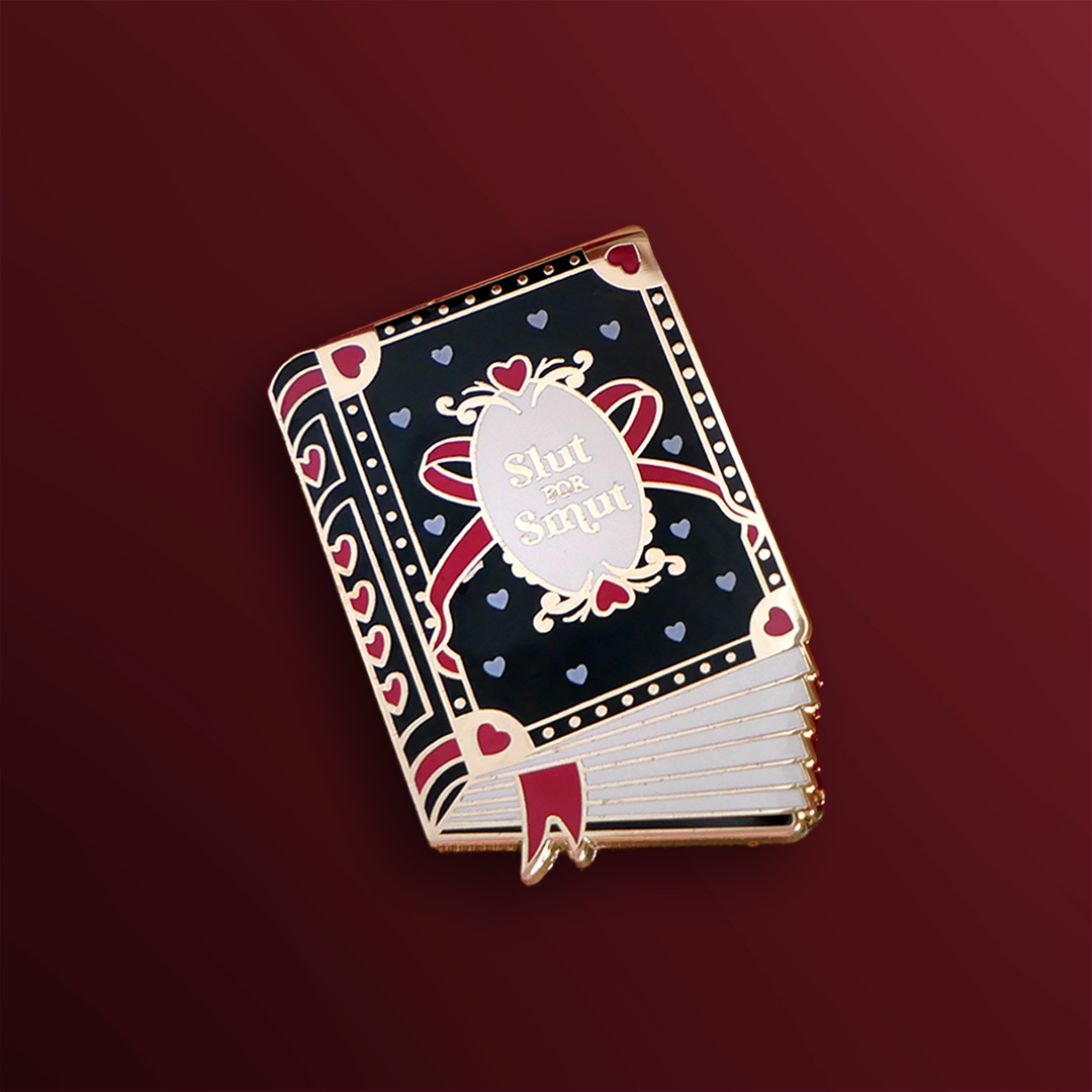 A an enamel pin in the shape of book that has a black cover with metallic with red and gold accents. The cover reads 'Slut for Smut' and features hearts and a bow