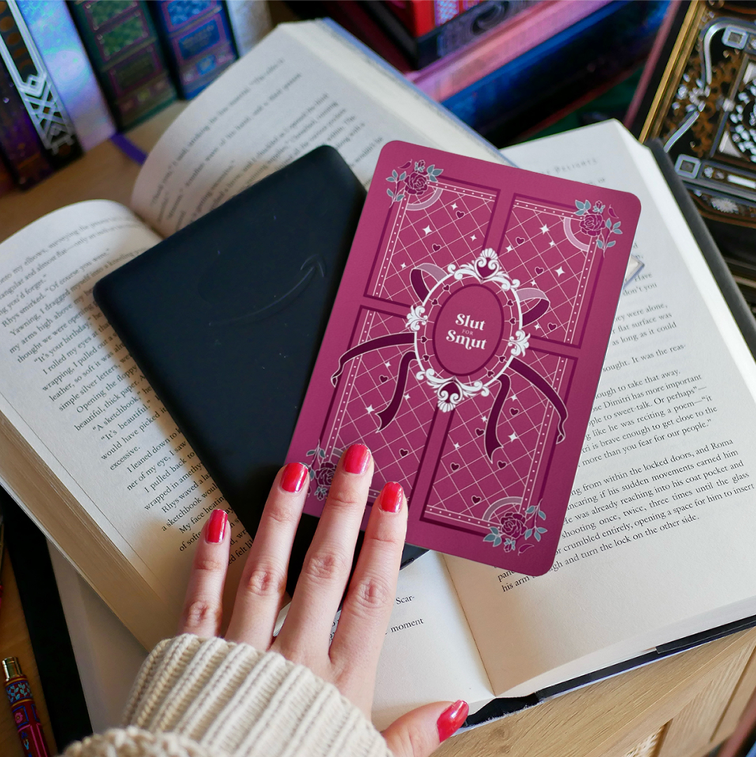 A hand holds the back of a kindle that sits on a bed of open books. On top of the kindle sits a sticker that reads 'slut for smut'. The sticker has pink, white and teal accents that features hearts, roses and a ribbon on a quilted style pattern