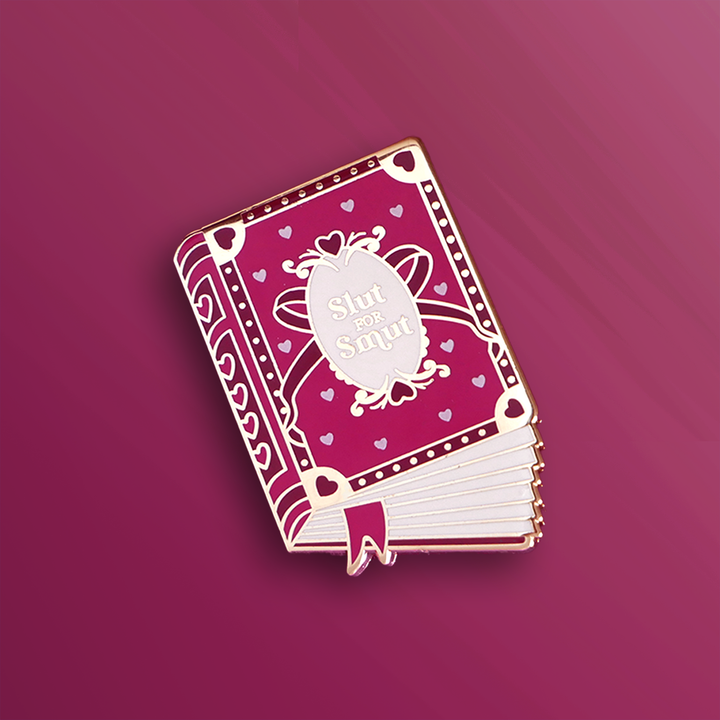 A an enamel pin in the shape of book that has a pink cover with metallic gold accents. The cover reads 'Slut for Smut' and features hearts and a bow