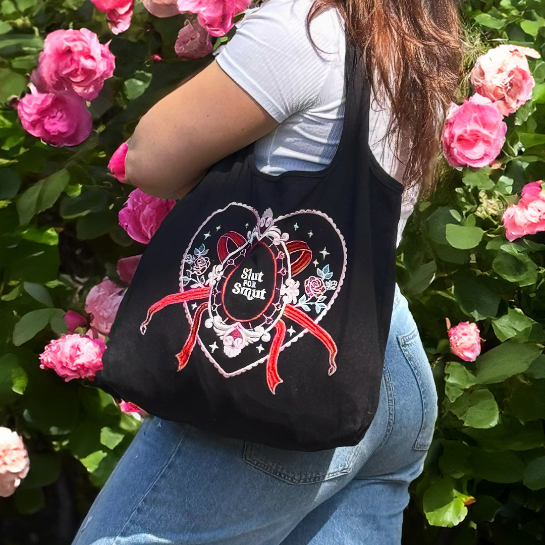 girl with brown hair, white top and blue jeans stands in front of a rose bush with pink flowers. Under her arm is a black tote bag with a design on the front that shows a heart with flowers, bows and stars. Phrase in the centre says 'slut for smut'