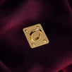 A The Quirky Cup Collective charm anchor on a dark red fabric background. The gold rectangle shaped clip is used to attach Charmlets to flat surfaces (such as your device).