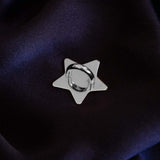 A The Quirky Cup Collective charm anchor on a dark purple fabric background. The silver star-shaped clip is used to attach Charmlets to flat surfaces (such as your device).