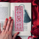 The Sugar & Spice bookmark is a light pink colour and features an intricate stained glass inspired design with motifs of birds, roses, hearts and leaves. The bookmark is held in front of a classical book on a red background and features a teal tassel.
