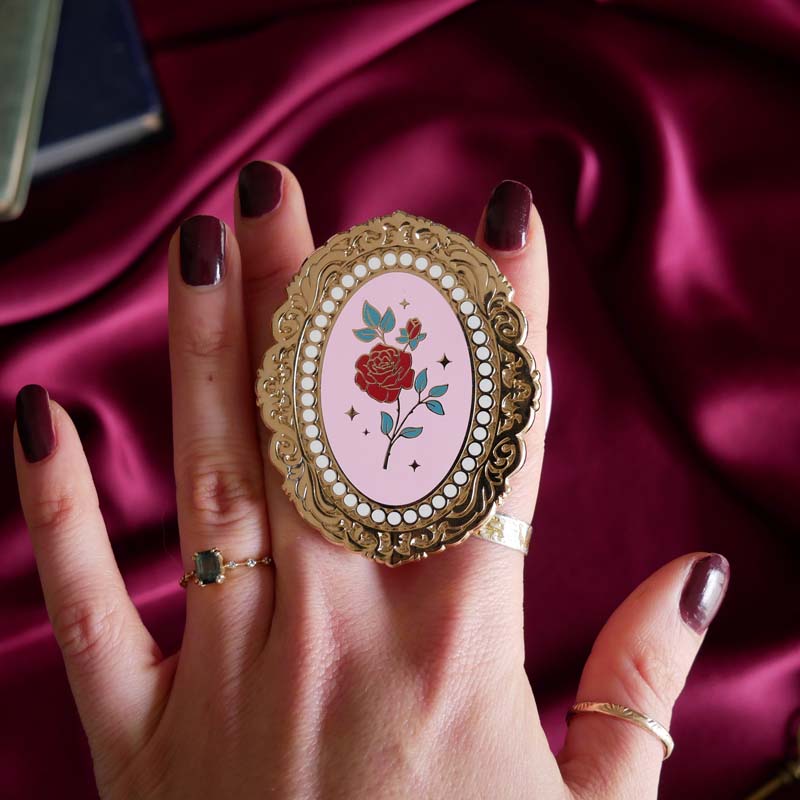 Hand holding a decorative kindle grip with a pink coquette rose inspired design with gold accents on a pink fabric background